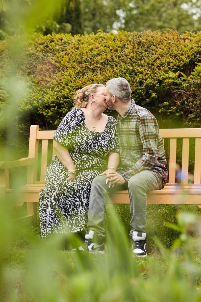 Engaged couple cuddling on a wooden bench, seen through foliage
