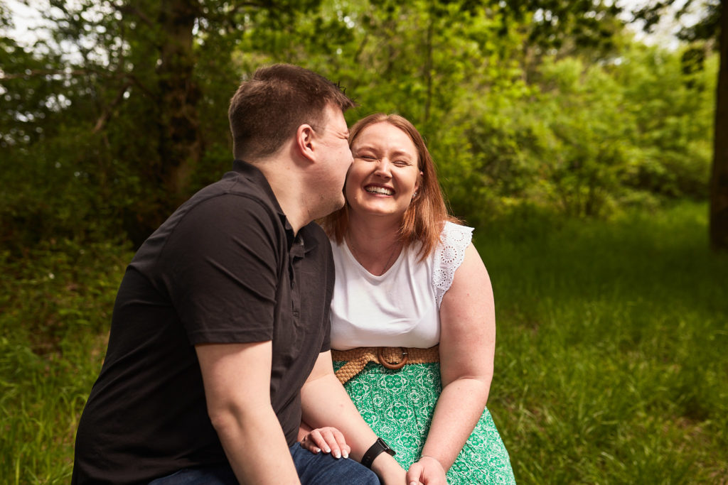 Man kissing his fiancée's cheek as she laughs in the woods