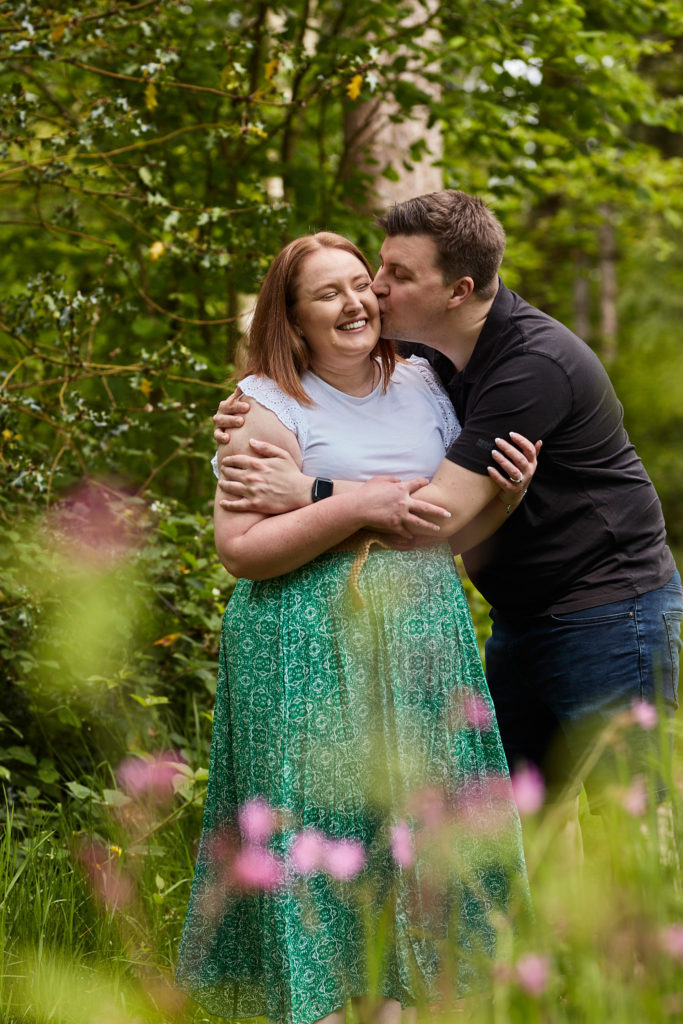 Man kissing his fiancée's cheek in the woods, pink wild flowers in the foreground
