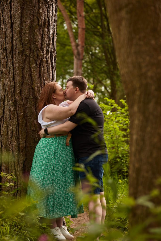 Engaged couple kissing between two pine trunks in the woods near Clacton