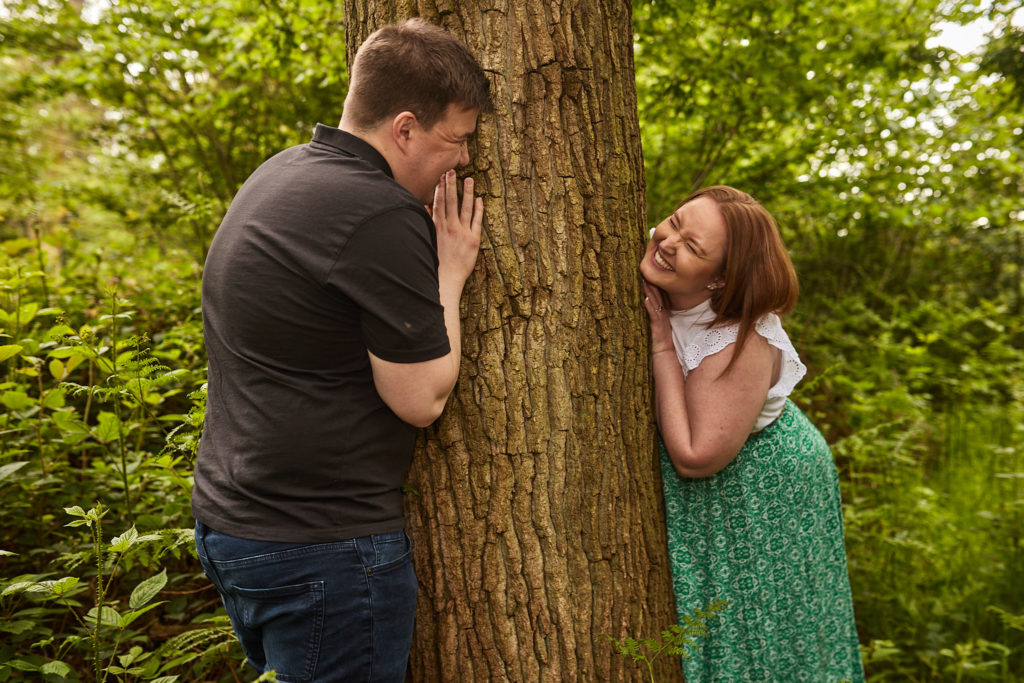 Engaged couple peeking round either side of a tree trunk at each other