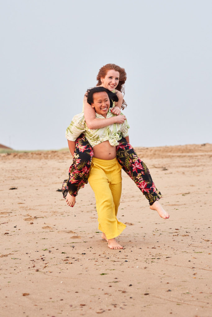 Woman laughing as she carries her fiancée on her back along the beach