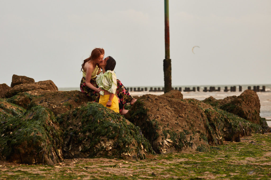 Engaged couple sitting on seaweed-covered rocks on the beach