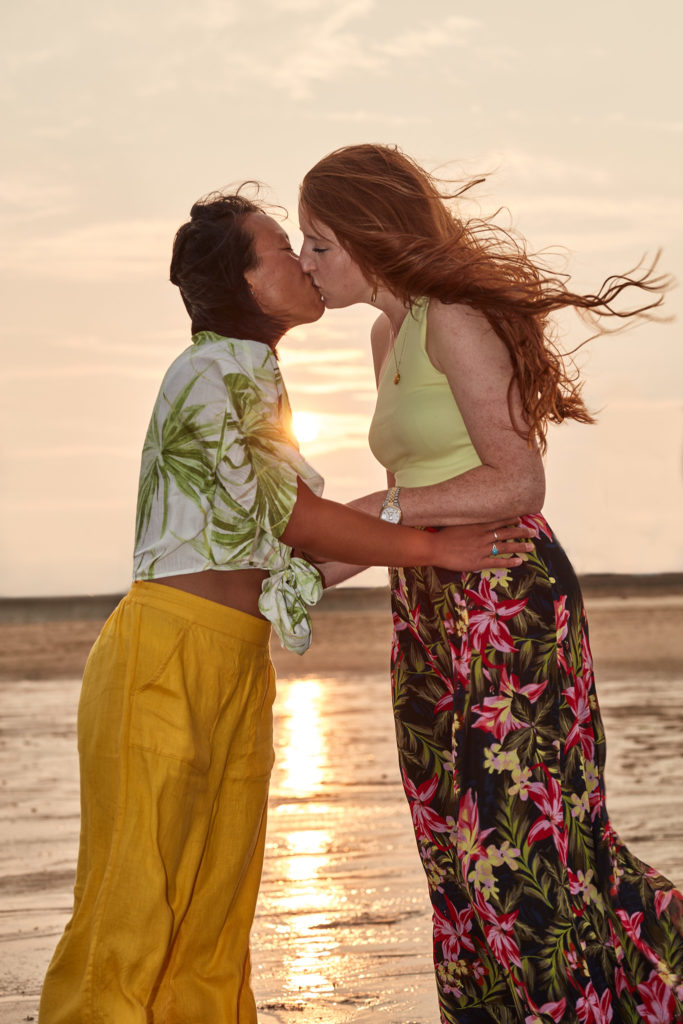 Two women kissing at the water's edge as the sun sets behind them