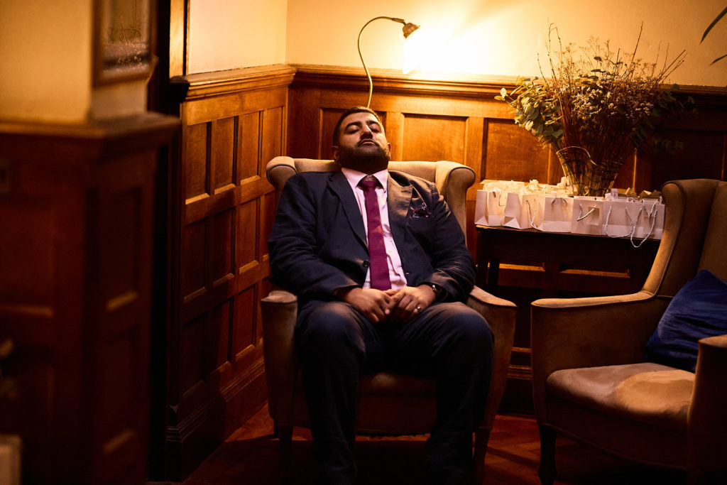 Groom in a navy suit sitting back in an armchair in a wood-panelled room at Prested Hall