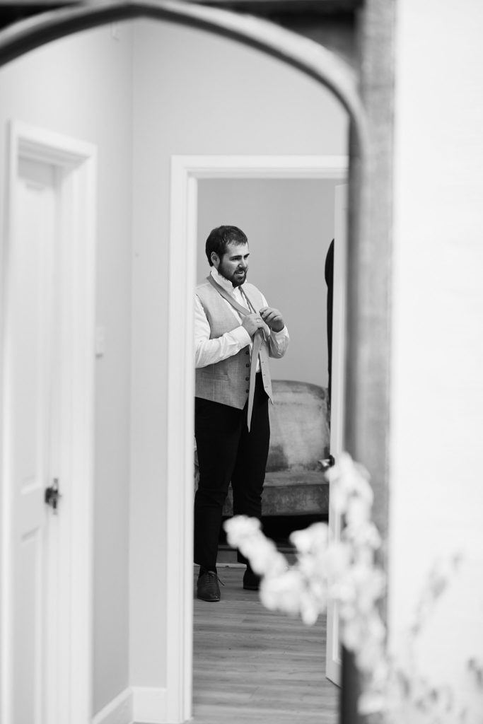 Black and white photo of the groom adjusting his waistcoat, seen through a doorway