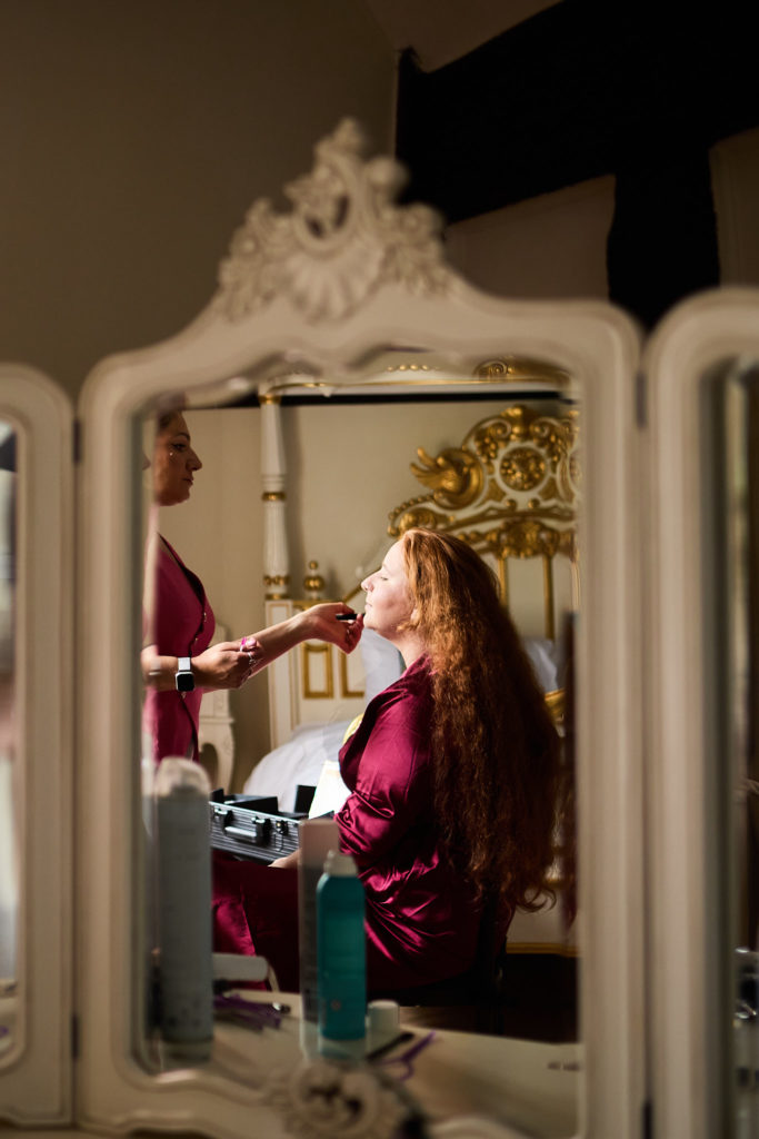 Bride in a burgundy robe having her make-up done, reflected in an ornate mirror