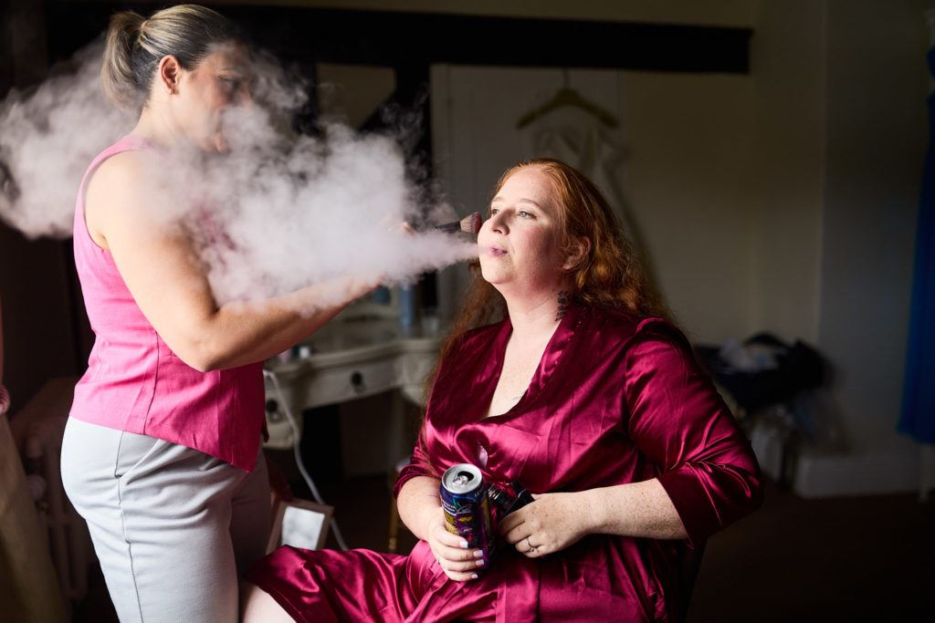 Hairdresser spraying a cloud of hairspray over the bride in a burgundy robe