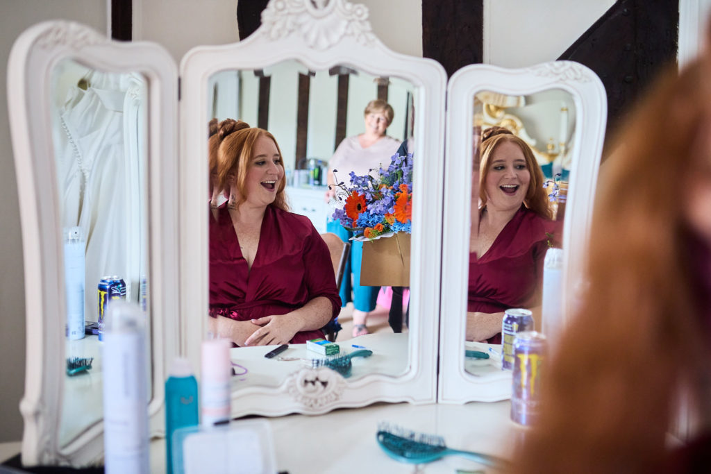 Bride and bridesmaid in burgundy robes laughing, reflected in a dressing table mirror