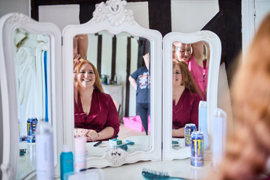 Bride and bridesmaid smiling at their reflections in a three-way dressing table mirror