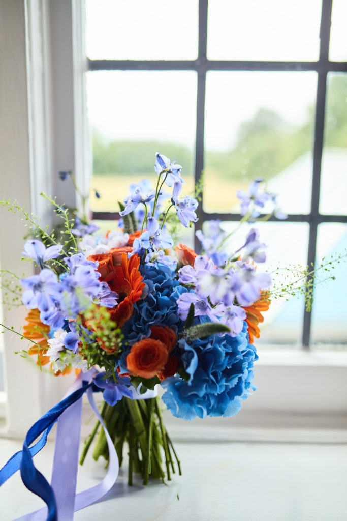 Bridal bouquet of blue, purple and orange flowers beside a leaded window