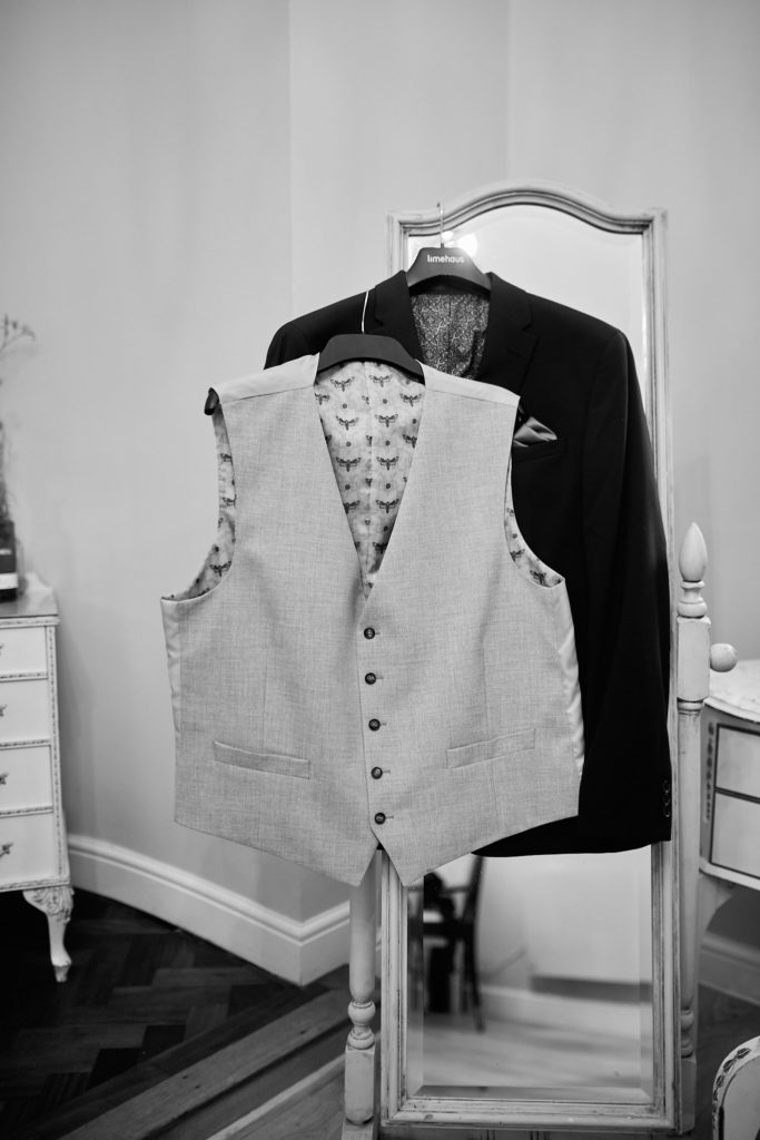 Black and white photo of the groom's waistcoat and jacket hanging on a mirror