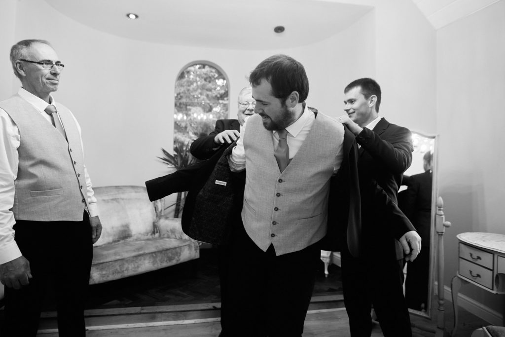 Black and white photo of groomsmen helping the groom into his jacket