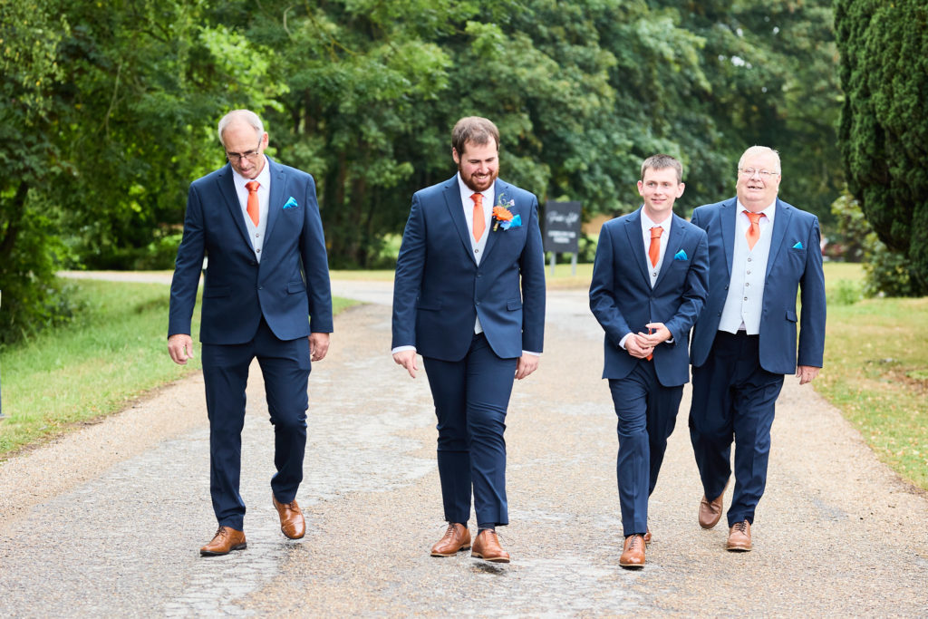 Groom and three groomsmen in navy suits walking along a tree-lined drive