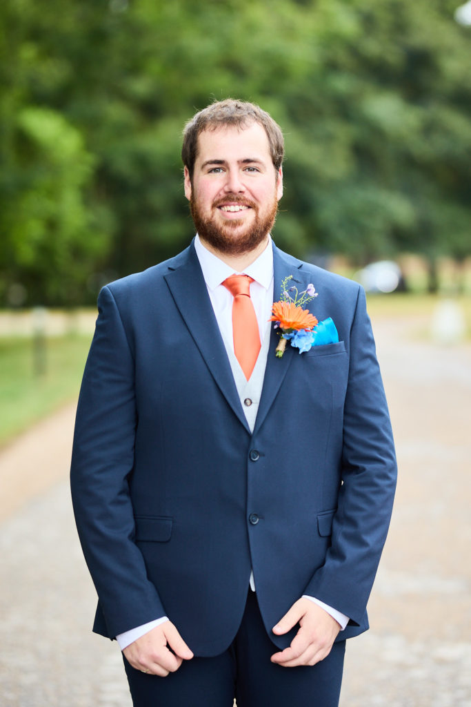 Groom smiling in a navy suit with an orange tie and buttonhole
