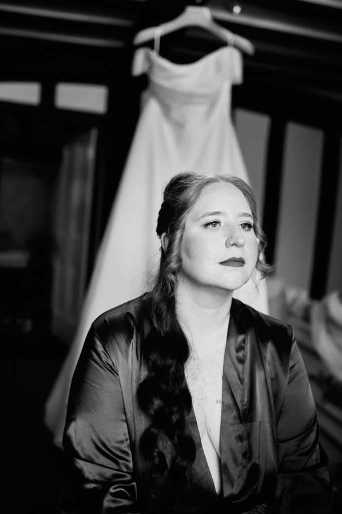 Black and white portrait of the bride in a satin robe with her wedding dress hanging behind