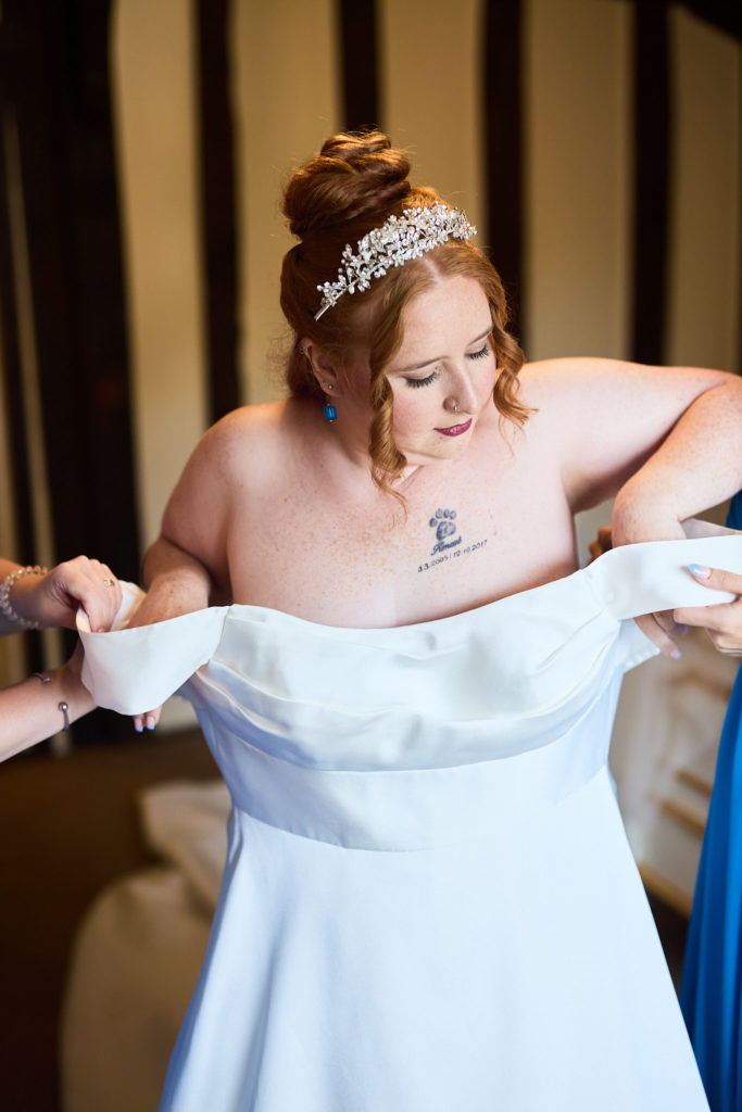 Bride in a tiara being helped into her off-the-shoulder wedding dress