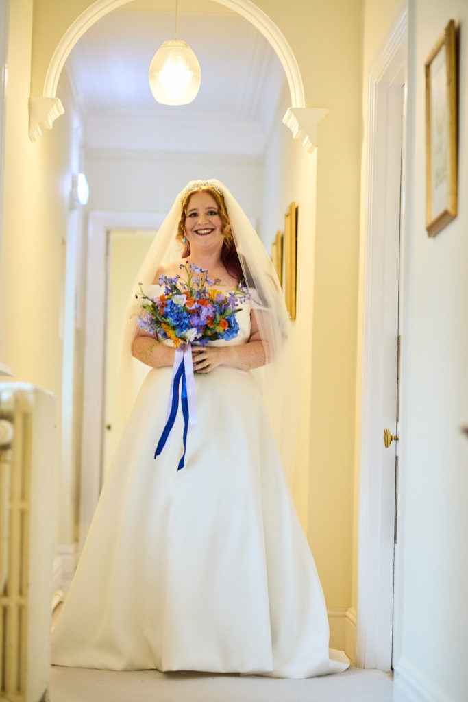 Bride in her veil holding a blue and orange bouquet in an arched hallway at Prested Hall