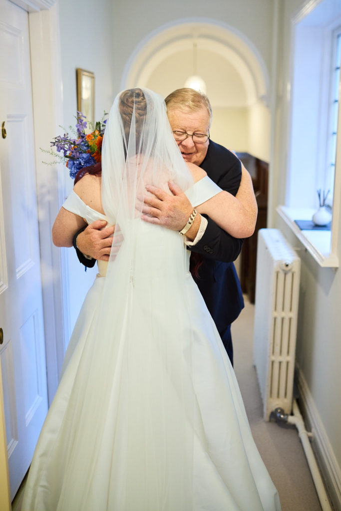 Father of the bride hugging his daughter in a hallway, her veil seen from behind