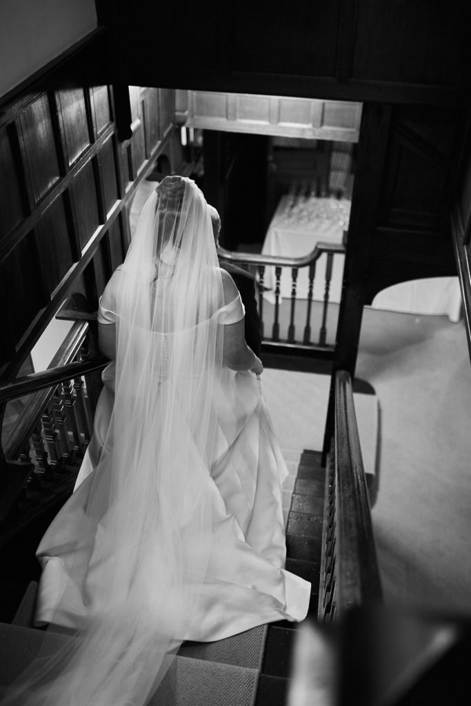 Black and white photo from above of the bride on a wood-panelled staircase with her long veil