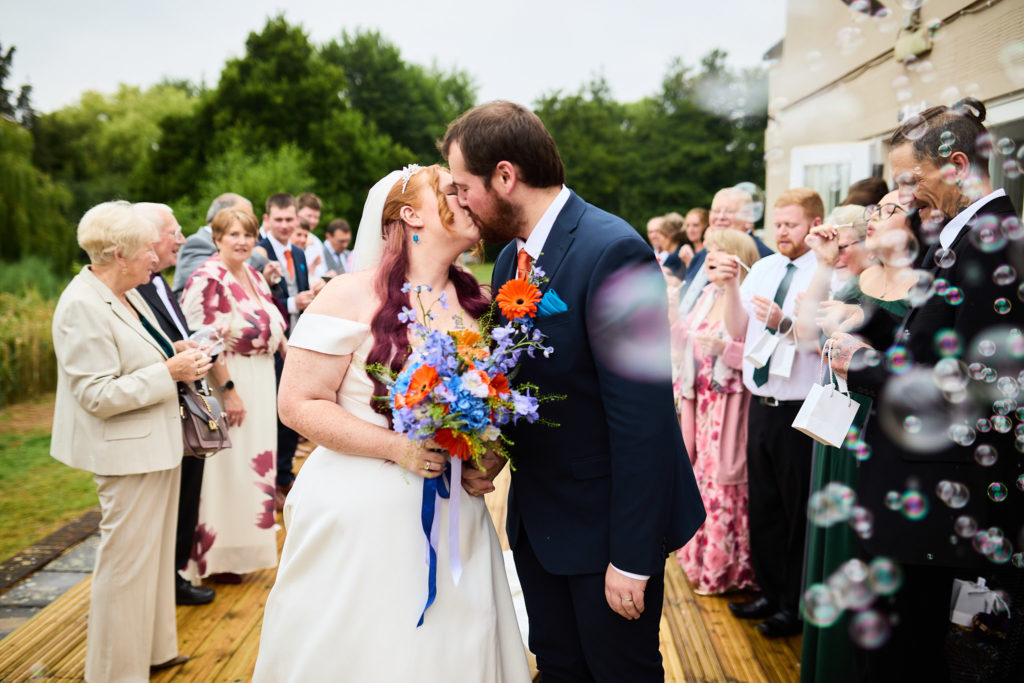 Bride and groom kissing as guests blow bubbles outside Prested Hall