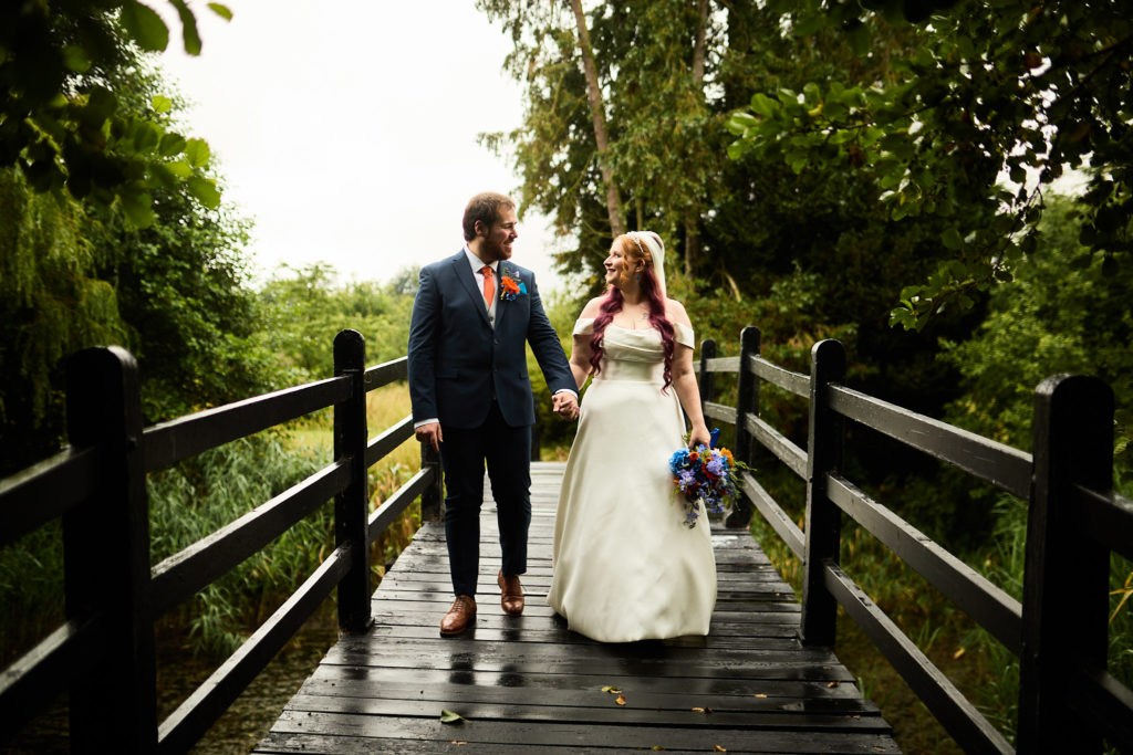 Bride and groom walking hand in hand across a wooden bridge at Prested Hall