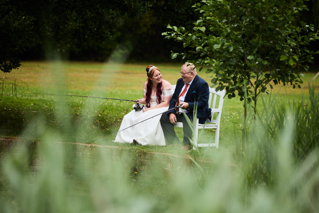 Bride and groom on a white bench in the grounds of Prested Hall, seen through long grass