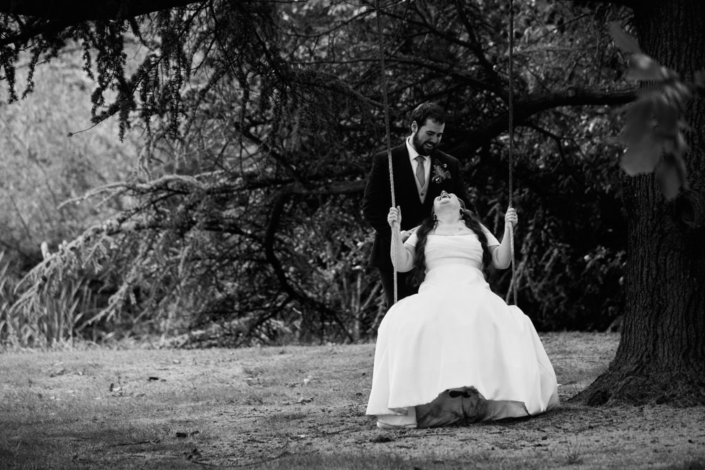 Black and white photo of the groom pushing the bride on a tree swing at Prested Hall