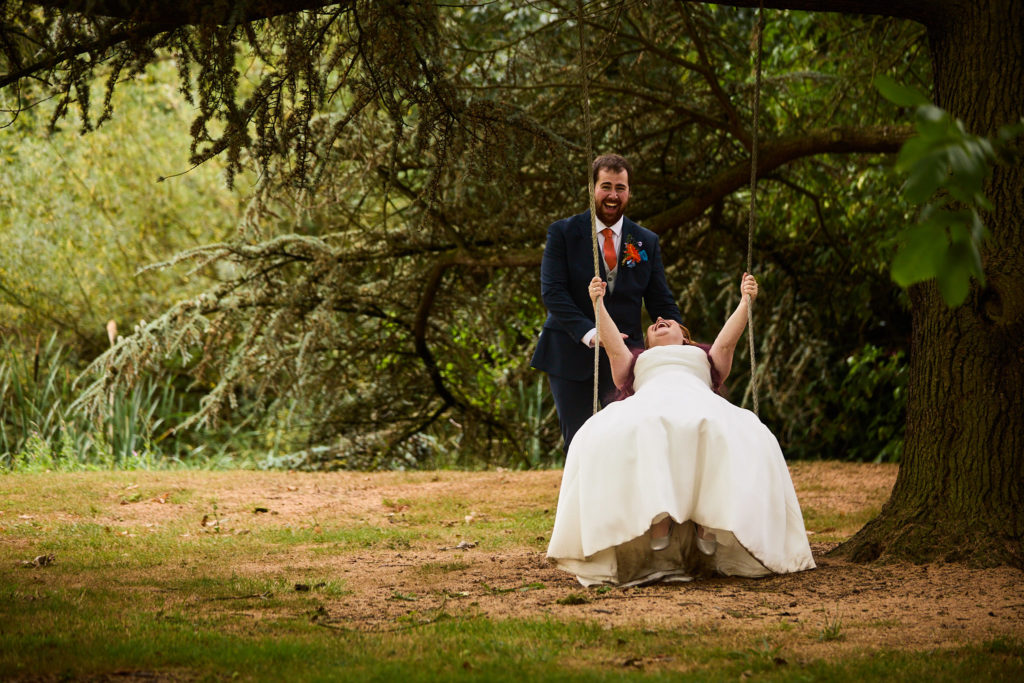Bride laughing on a tree swing with the groom behind her in the grounds of Prested Hall