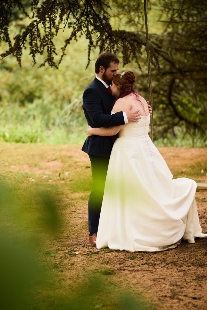 Groom kissing the bride's forehead as they hug under a tree at Prested Hall, Colchester