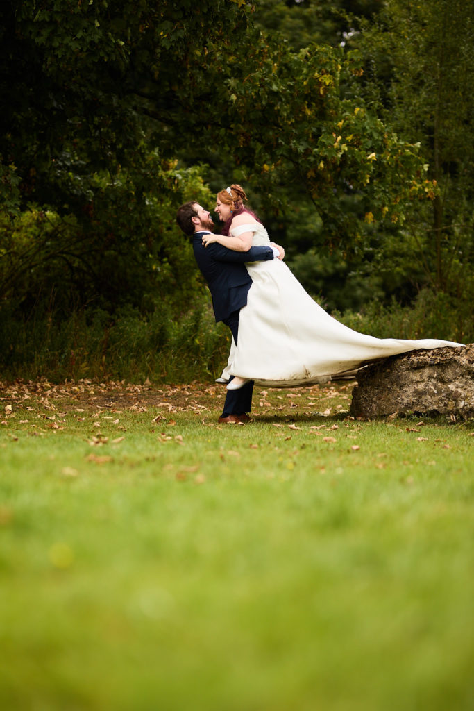Groom holding the bride in his arms beside a rock in the Prested Hall gardens, her dress train flowing