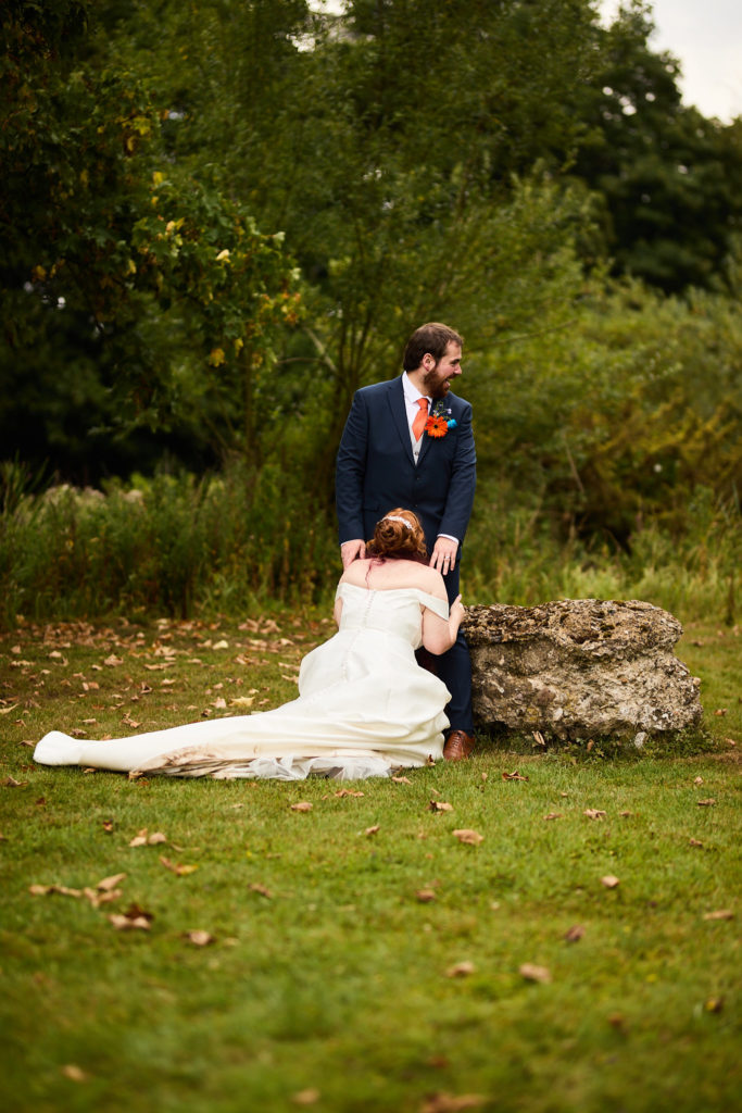 Bride sitting on the grass beside a rock with the groom standing over her at Prested Hall