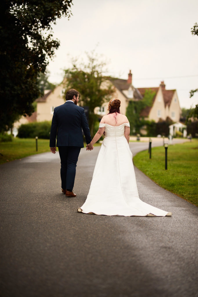 Bride and groom walking hand in hand towards Prested Hall, seen from behind