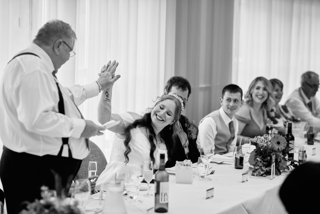 Black and white photo of the father of the bride giving his speech as the bride laughs at the top table