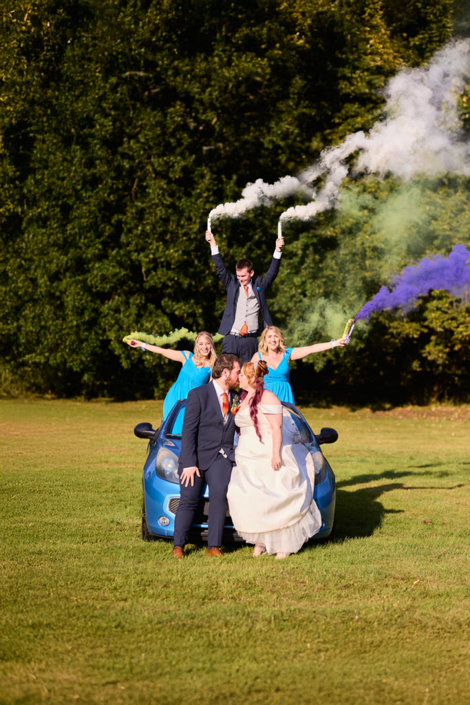 Bride and groom kissing beside a blue convertible as the bridal party wave smoke flares