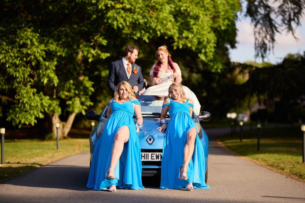 Bride and groom sitting on a blue convertible with bridesmaids in blue dresses in front