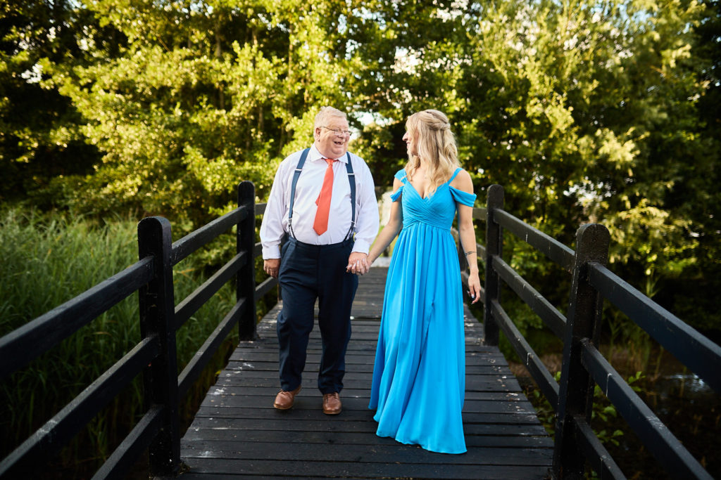 Father of the bride and a bridesmaid in a blue dress walking across a wooden bridge