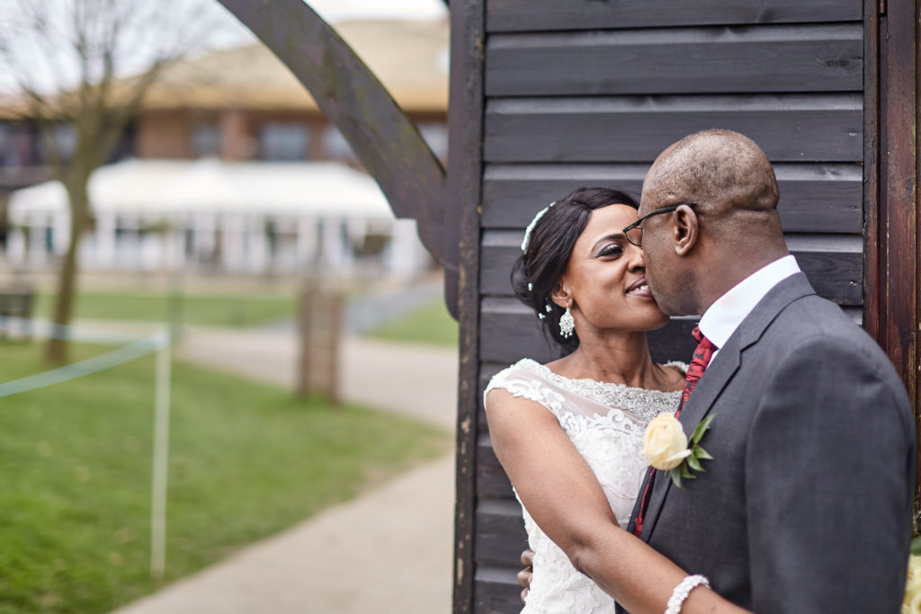 Groom kissing the bride's cheek beside a black timber building at Essex Golf Club