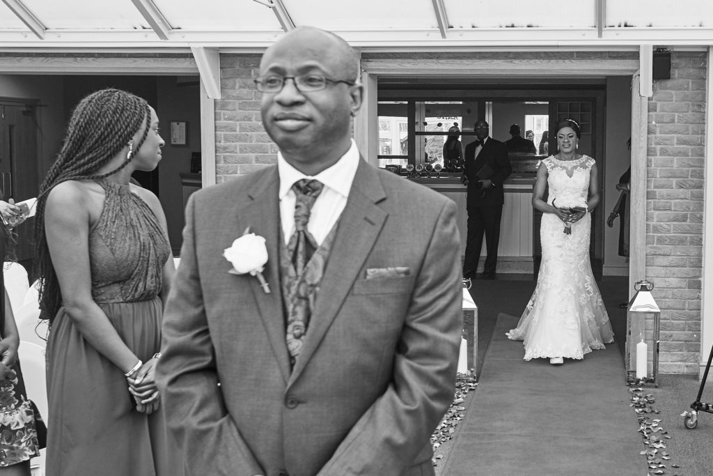 Black and white photo of the groom waiting as the bride arrives behind him