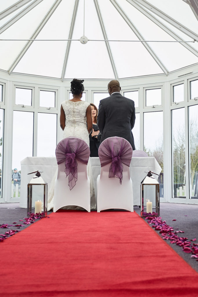 Bride and groom standing at the front of the conservatory ceremony room, seen from the aisle
