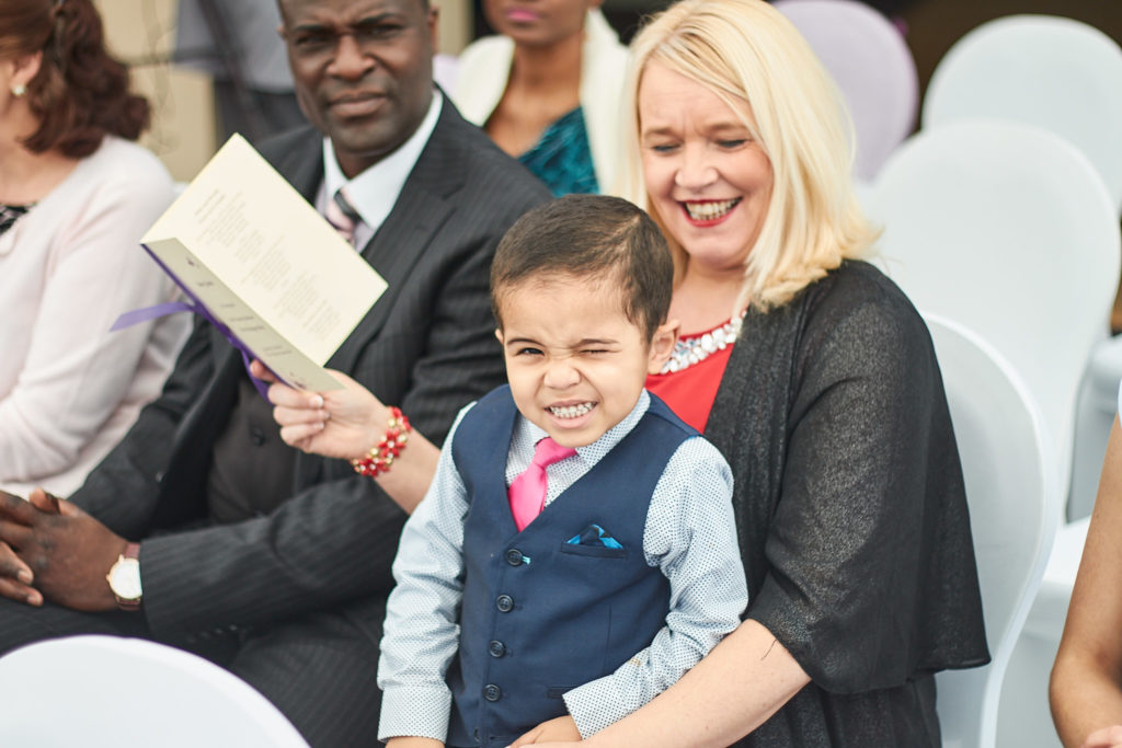 Young boy in a waistcoat pulling a face on a guest's lap during the ceremony