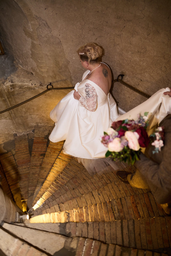 Bride descending a spiral stone staircase with her bouquet at Hedingham Castle, seen from above