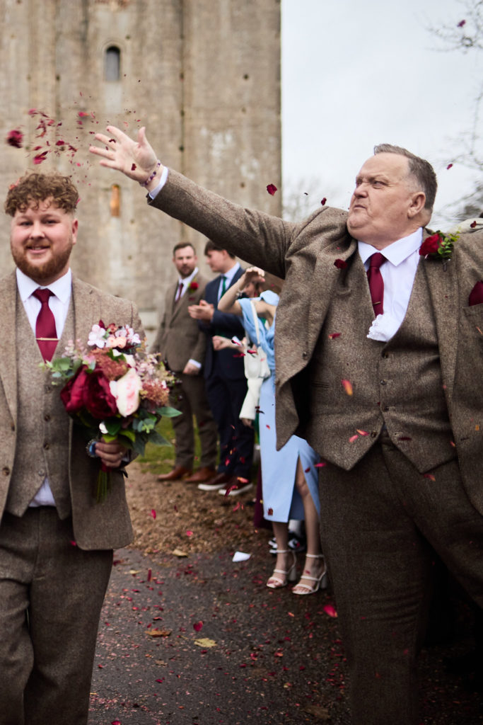 Wedding guest in a tweed suit throwing confetti as the groom laughs