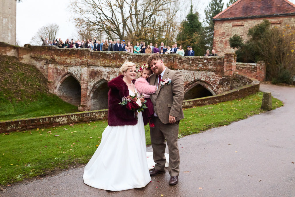 Bride and groom in front of the Tudor brick bridge at Hedingham Castle with guests lined up along it