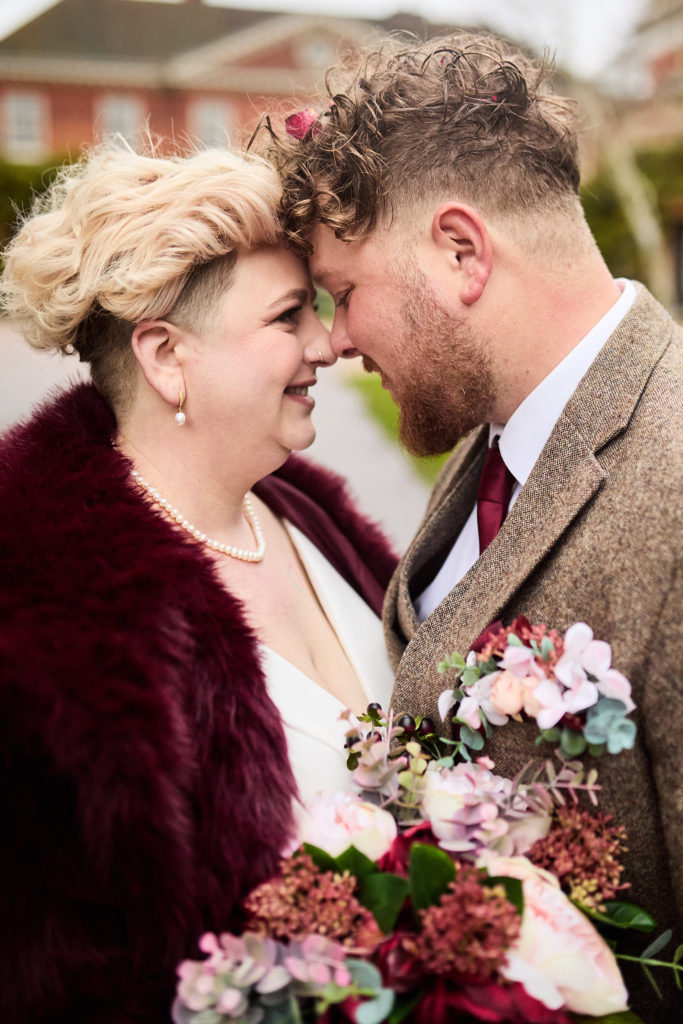 Bride in a burgundy fur jacket laughing forehead to forehead with the groom at Hedingham Castle