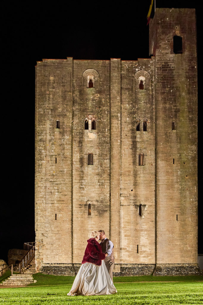 Bride and groom kissing in front of the floodlit keep at Hedingham Castle at night