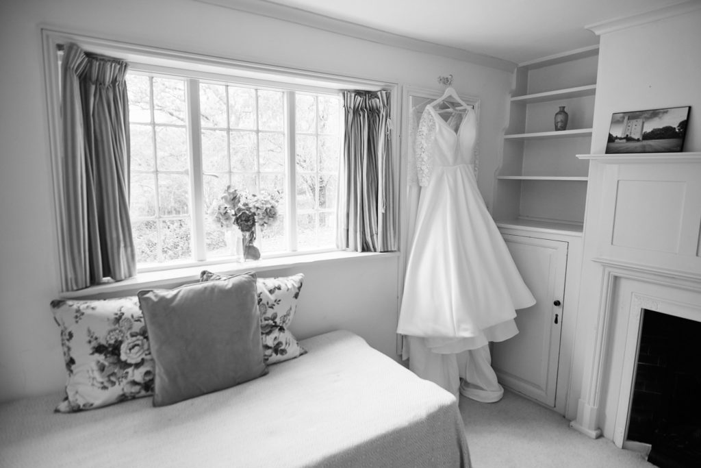 Black and white photo of the wedding dress hanging in a bedroom beside a window
