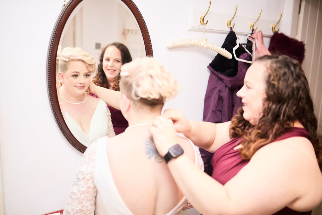 Bridesmaid fastening the back of the bride's dress, reflected in an oval mirror