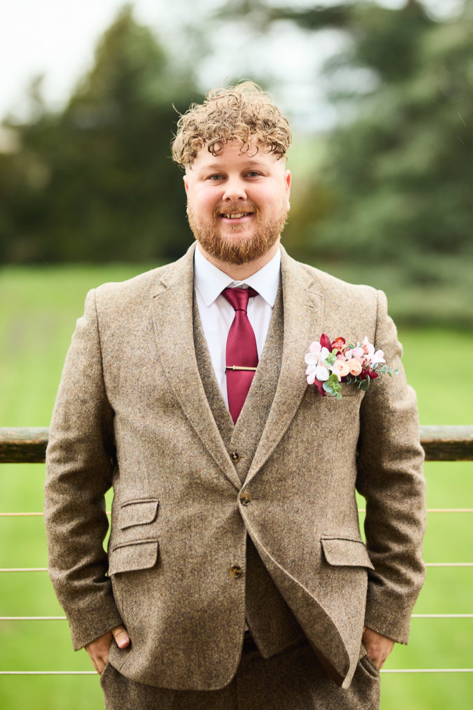 Groom smiling in a tweed suit with a burgundy tie and buttonhole