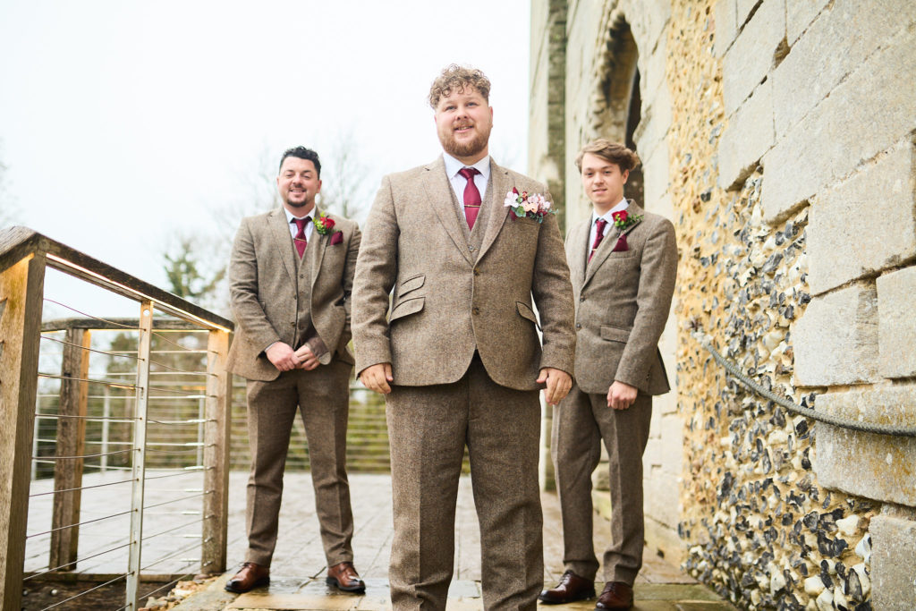 Groom and two groomsmen in tweed suits outside Hedingham Castle